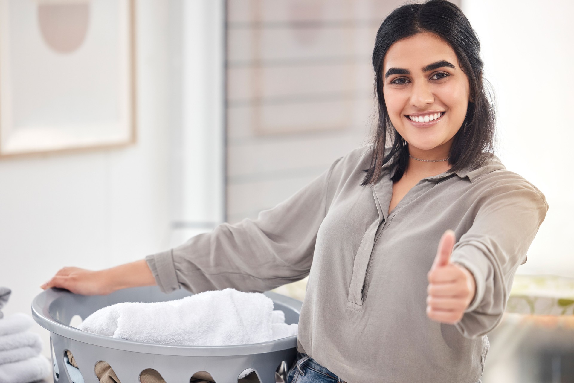 Shot of a young woman showing a thumbs up while doing laundry at home